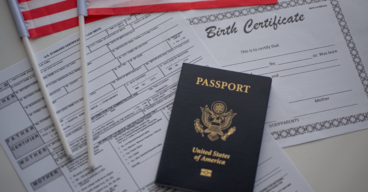 A black U.S. passport lies on birth certificate and application papers beside small American flags on a table.