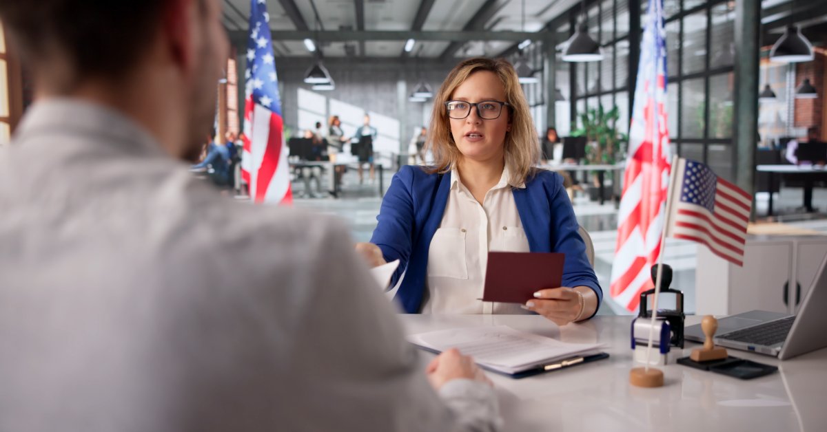 A woman with glasses holds a red booklet across a desk from another person, with U.S. flags and a laptop nearby.