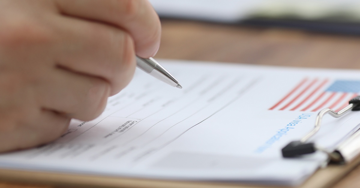 A hand holding a pen over an application document on a clipboard, with a small U.S. flag printed on the page.