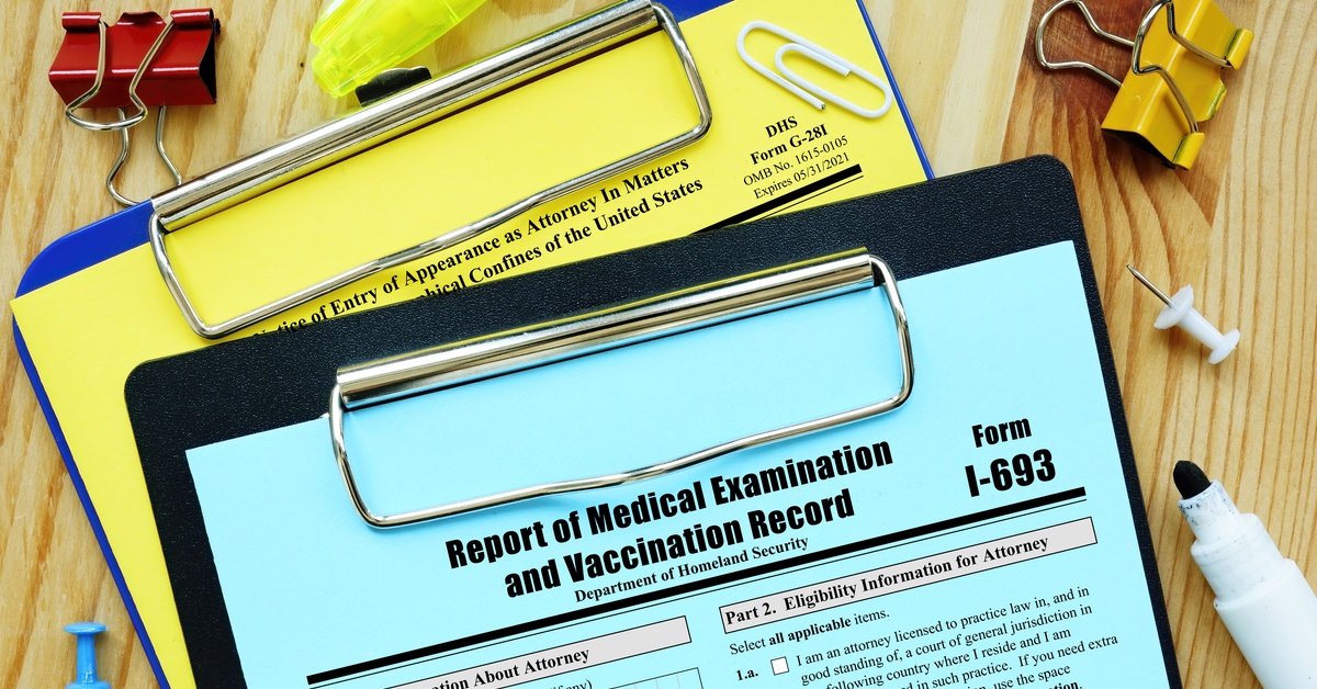 A blue Form I-693 document on a clipboard with binder clips, paper clips, push pins, and a highlighter on wood.