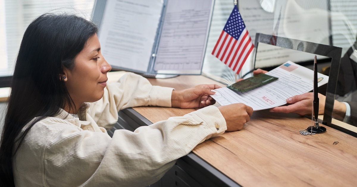 A woman passing a passport and paperwork across a desk to hands behind a clear partition, with a U.S. flag nearby.