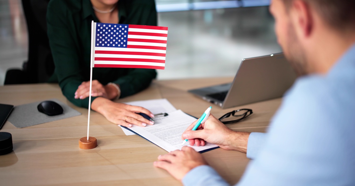 A person writing on a document at a desk with a small United States flag between two seated people indoors.