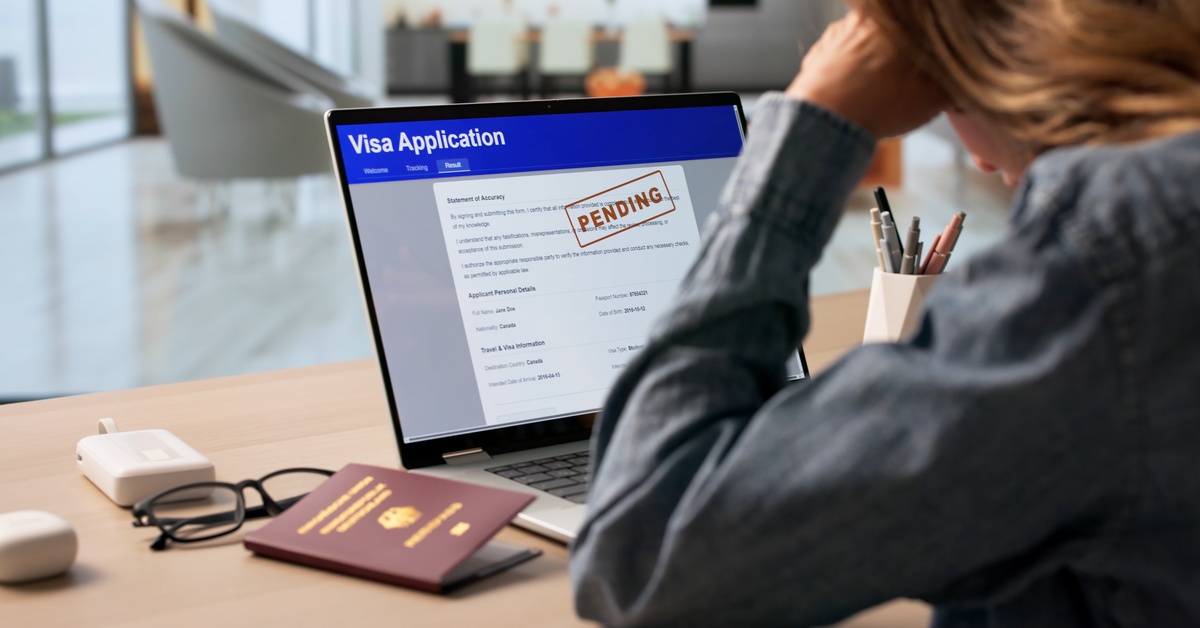 A woman seated at a desk with a passport, glasses, and a laptop displaying a pending visa application.
