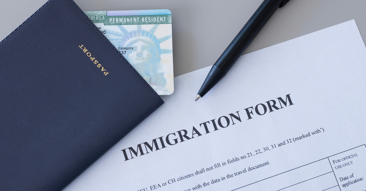 A blue passport and permanent resident card resting beside a black pen and immigration form on a gray desk.