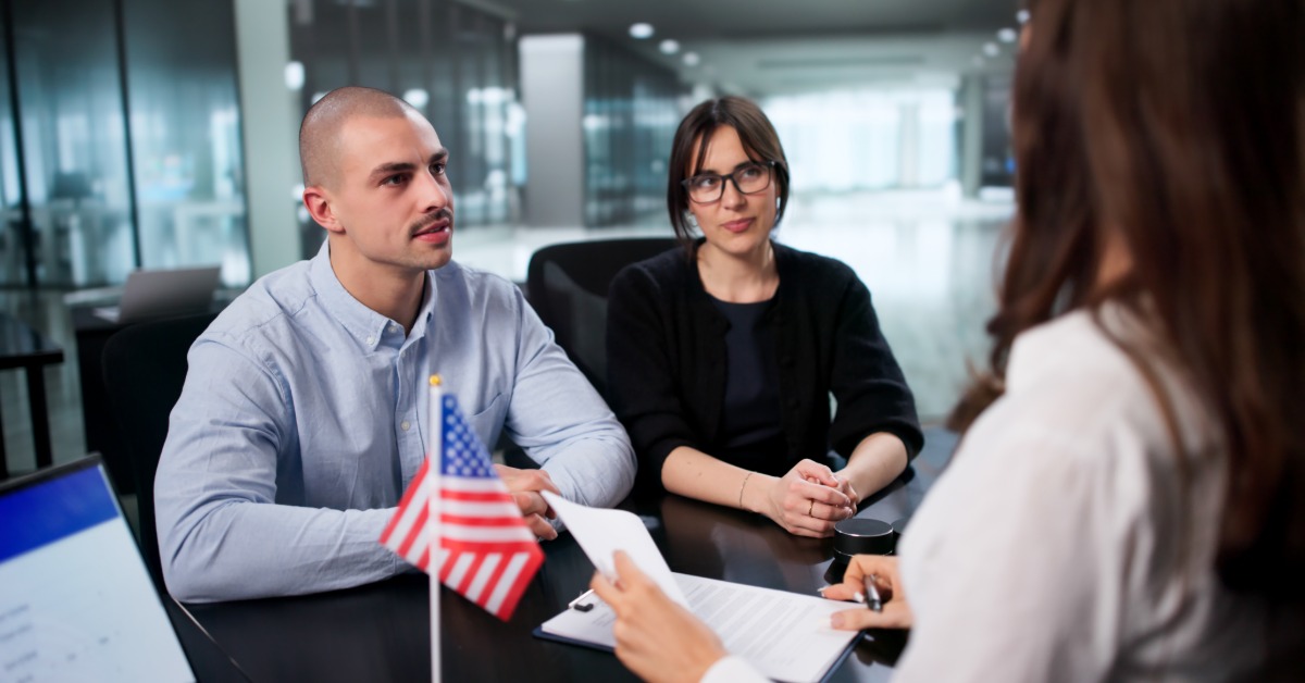 Two people sitting across from a woman holding papers at a desk with a small United States flag nearby.