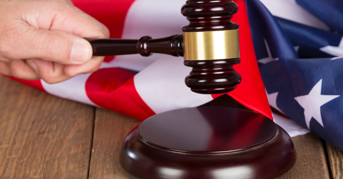 A hand holding a wooden gavel above a round sound block on a table, with a folded American flag behind it.