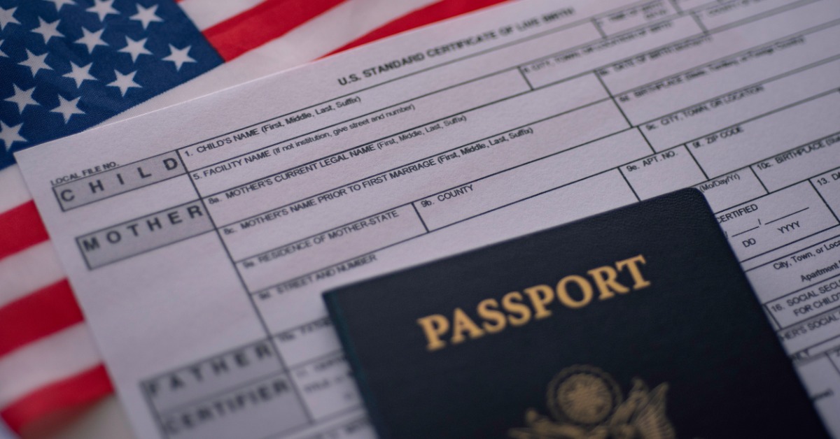 A dark blue U.S. passport resting on a standard certificate of live birth form beside a partially visible American flag.