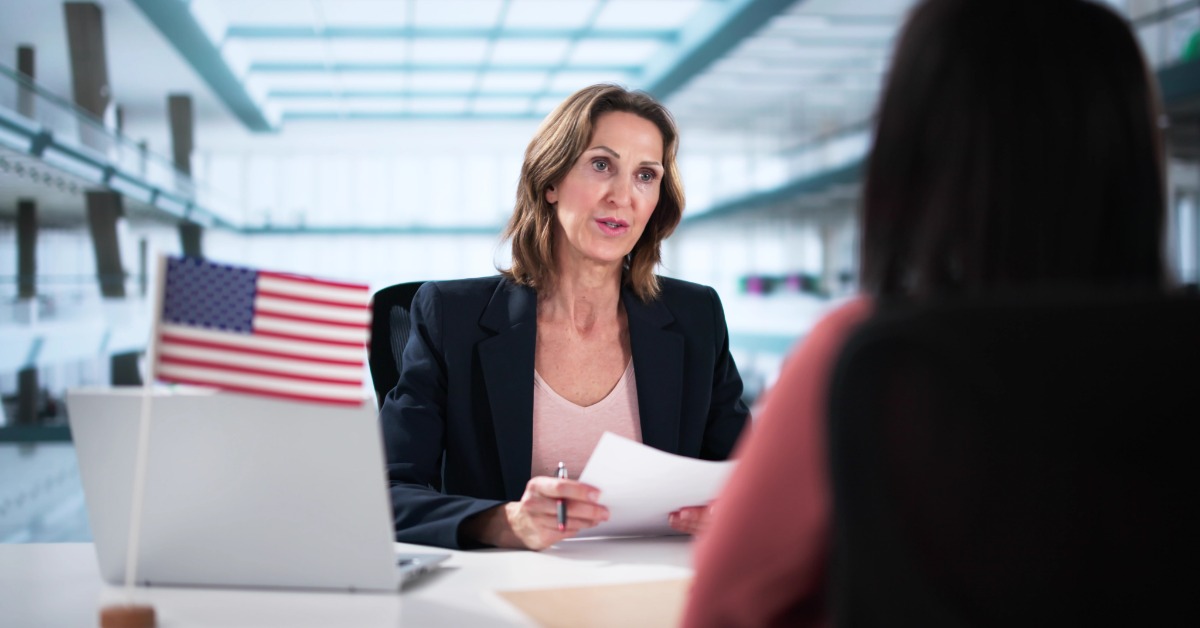 A woman in a dark blazer holding papers across a desk from another woman beside a laptop and a small American flag.