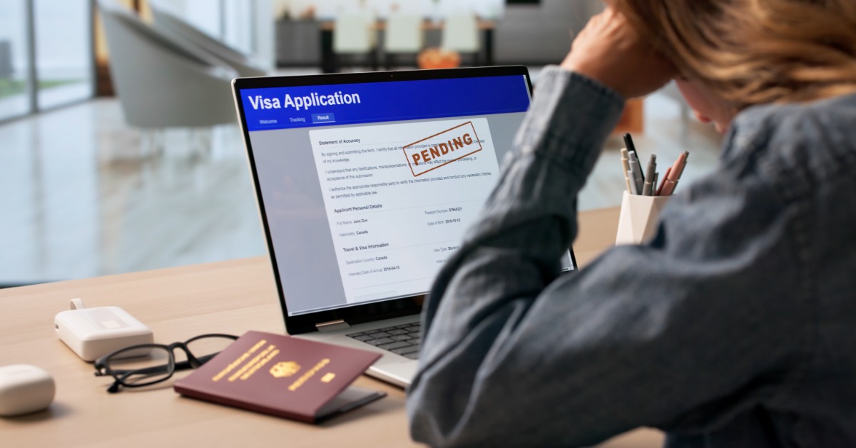 A woman leaning toward a laptop showing a visa application marked pending, with a passport and eyeglasses on the desk.