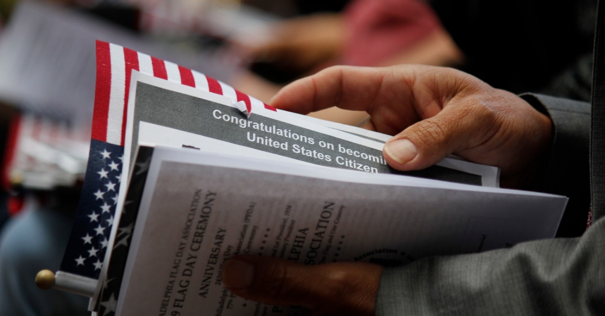 A person's hands holding a stack of citizenship documents with a small American flag and blurred people in the background.