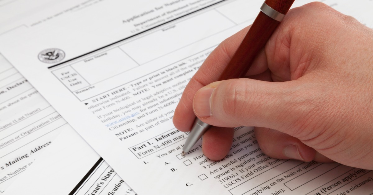 A hand holding a brown pen over a checkbox on a U.S. naturalization application with other paperwork underneath.