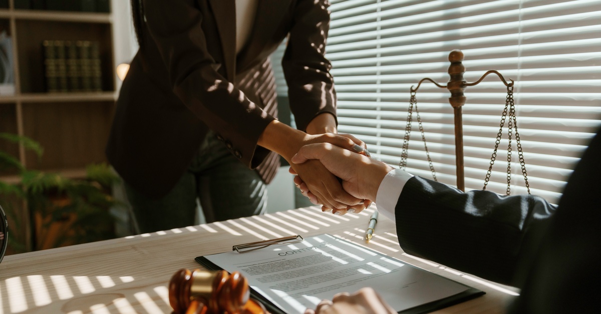 A person is standing up and shaking hands with another person who is sitting down. There is a clipboard with a slip of paper between them.