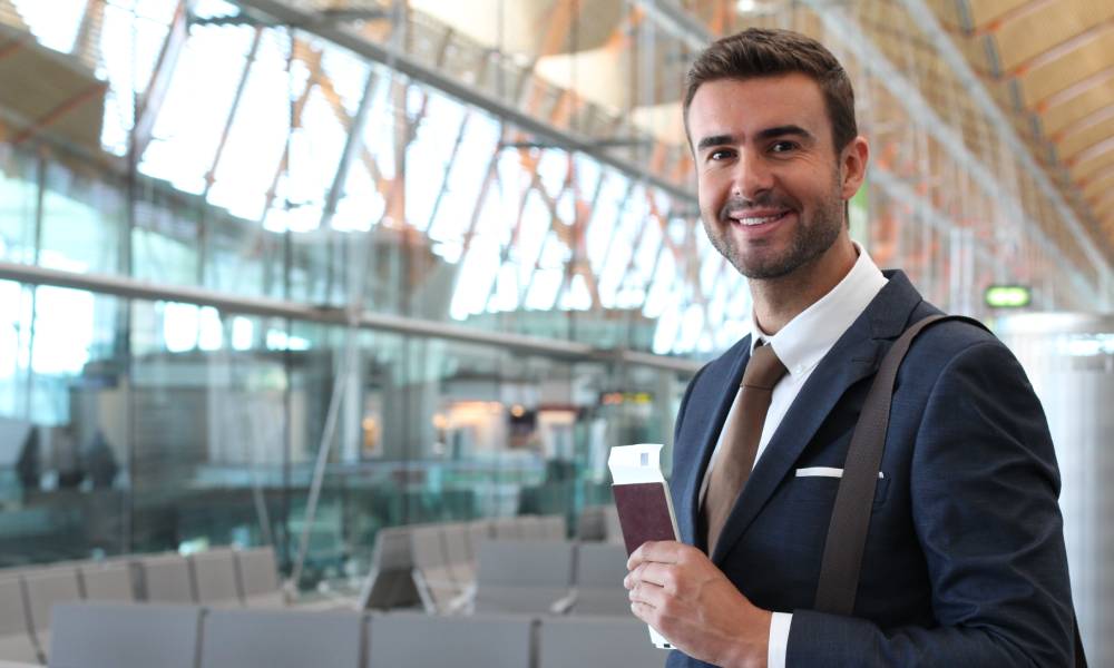 A smiling businessman dressed in a suit standing in the airport. He is holding his passport and boarding pass.