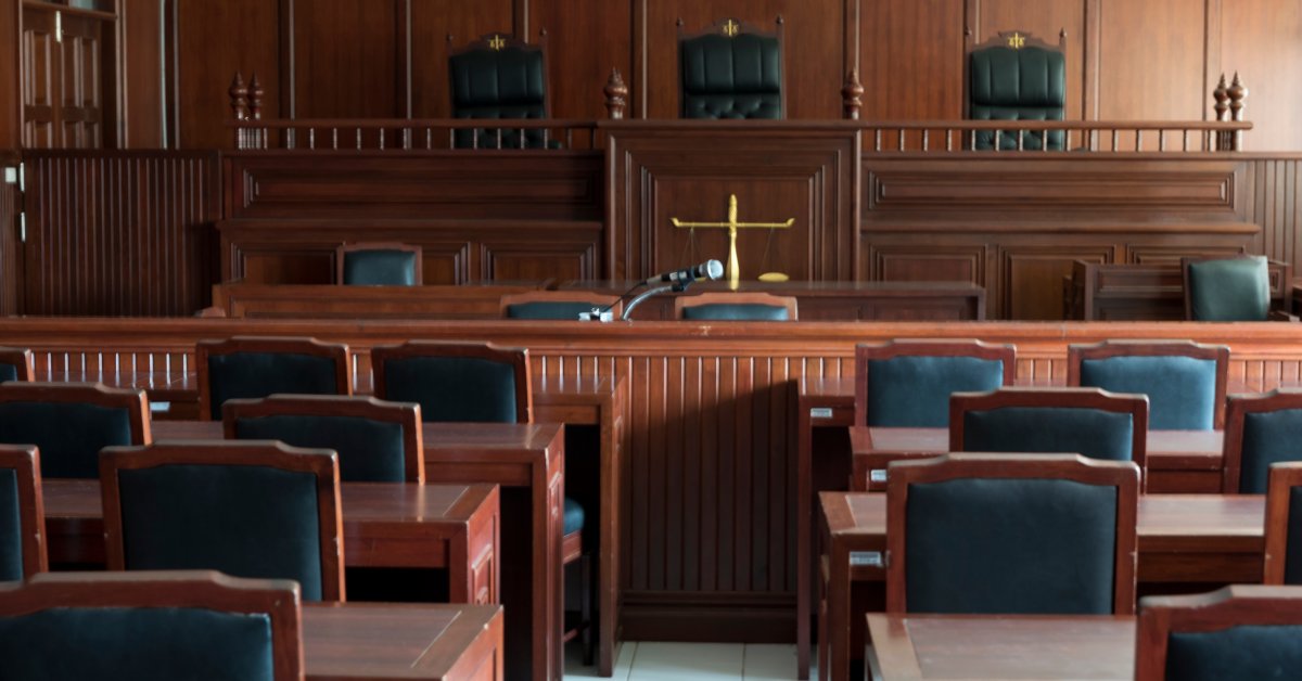 The interior view of an empty courtroom. There are several wooden tables and chairs facing the judge's bench.