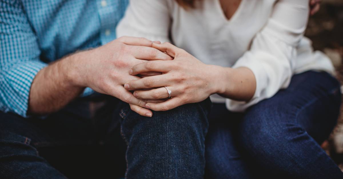 A man and a woman sitting close together and holding hands. The woman is wearing a diamond ring on her left ring finger.