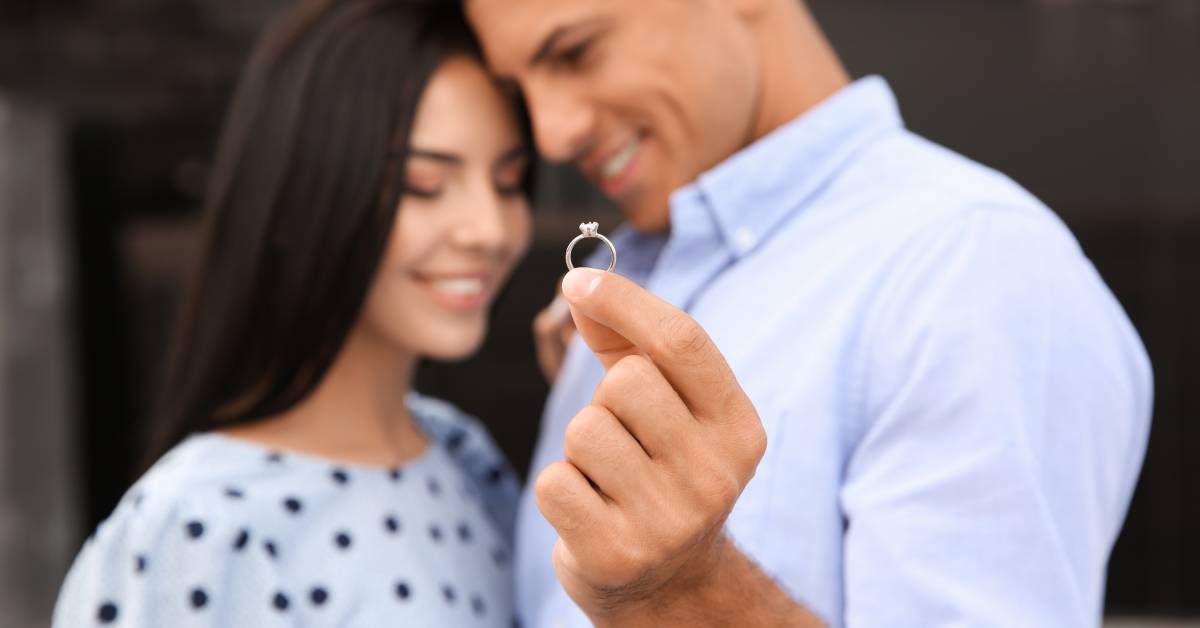 A man and a woman standing next to each other, touching foreheads, and smiling. The man holds up a diamond ring.