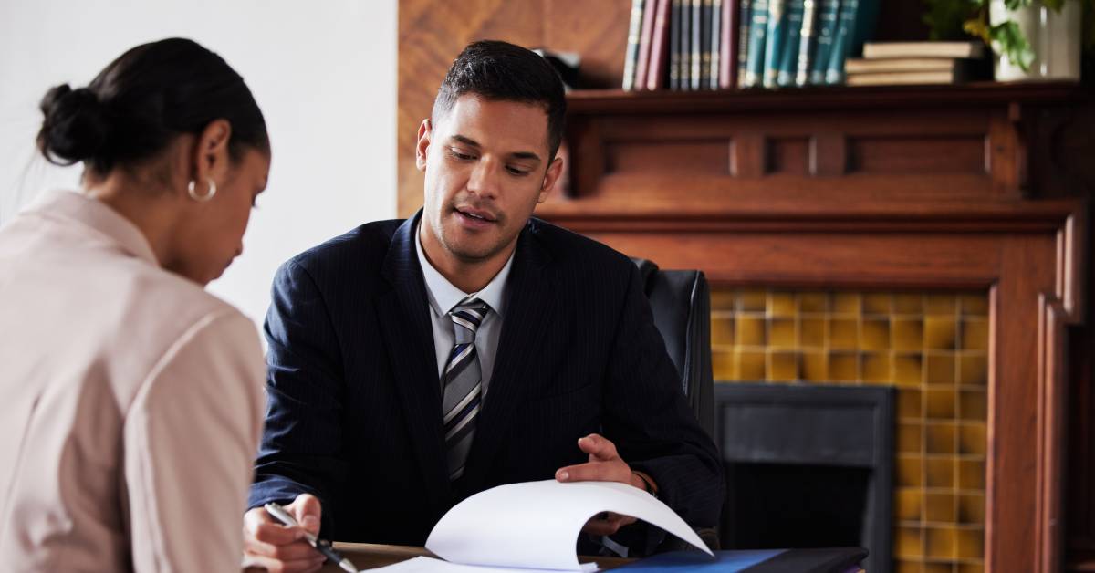 A man is wearing a suit and sitting across from a woman wearing a blazer. He is explaining paperwork with a pen in his hand.