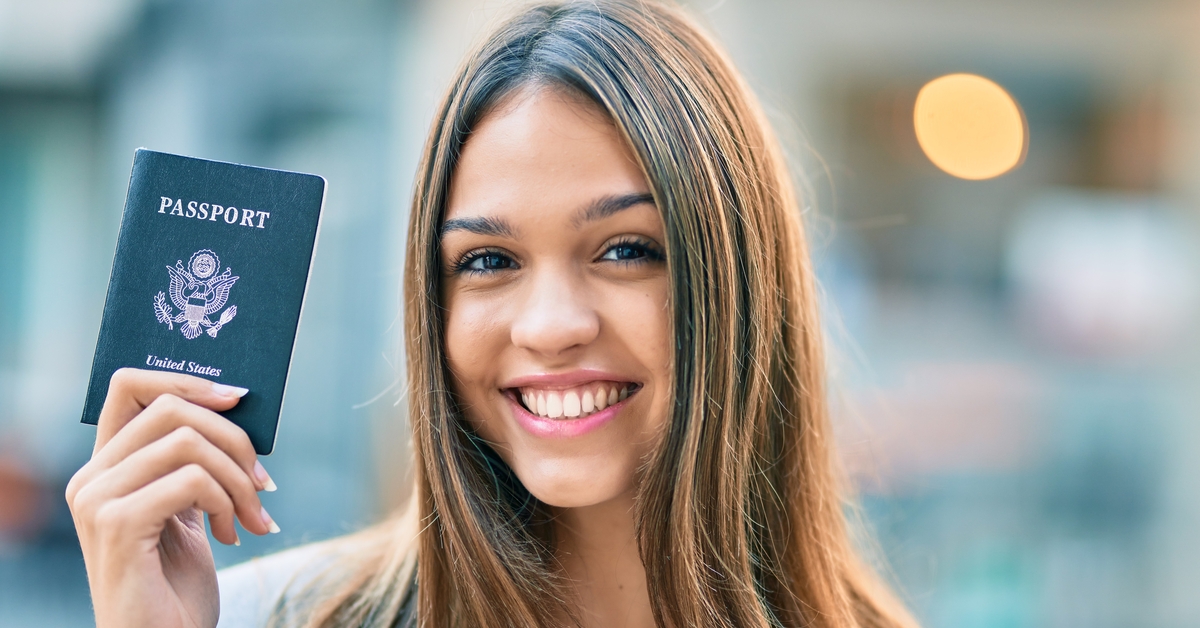 A woman wearing professional clothing is smiling and holding up a United States passport book in her right hand.