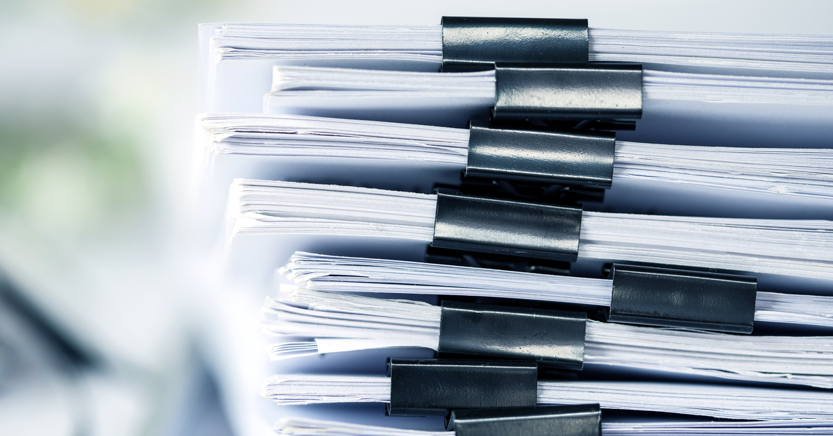 A stack of white documents organized by black paper clips. A pencil and a pair of glasses sit next to the papers.