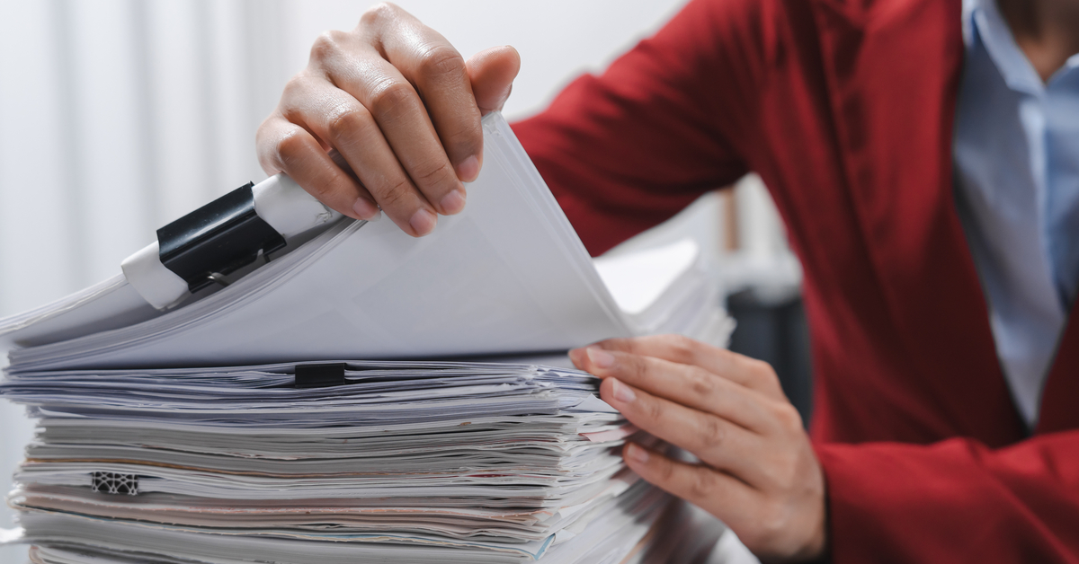 An individual wearing a red blazer, sitting at a desk and sorting through a very large stack of paperwork.