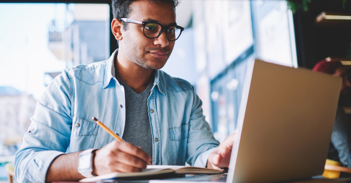 A man wearing dark glasses and looking at his laptop. He is using a pencil to write notes in a notebook.