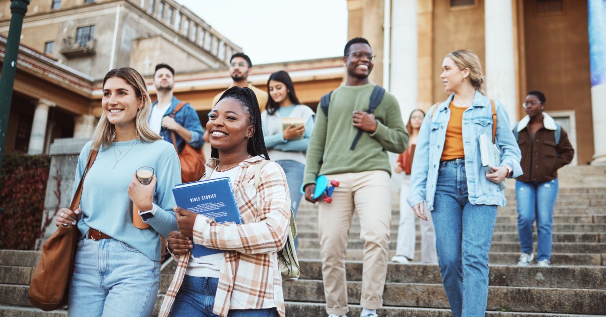 A group of students walking down stairs in front of a large building. They are wearing backpacks and holding books.