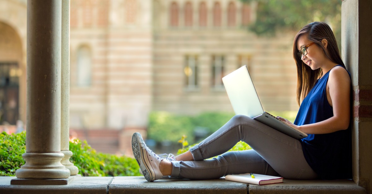 A young woman sitting outside and looking at her laptop while smiling. She has a book and pen next to her.