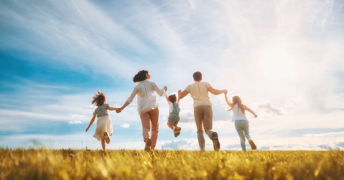 A rear view of a mother, father, and three children holding hands. They are walking in an open field and the sun is shining.