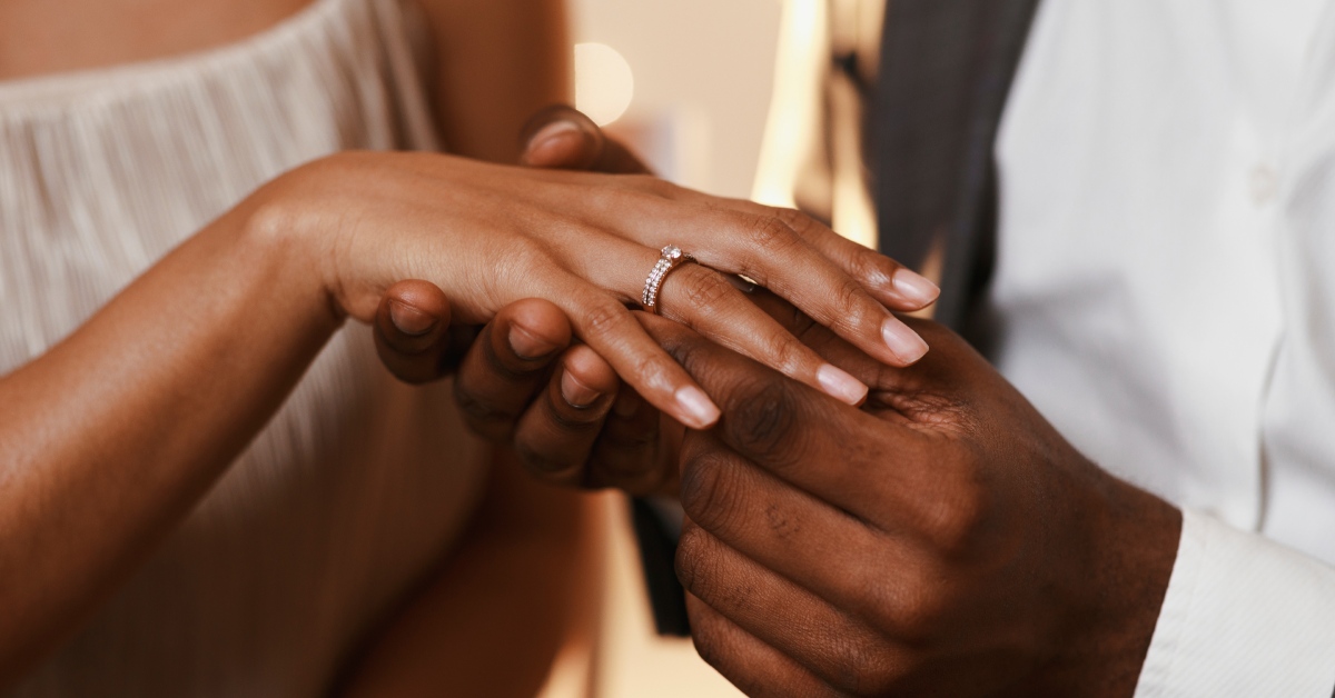 A man and a woman standing close together. The man is putting a diamond ring on the woman's ring finger.