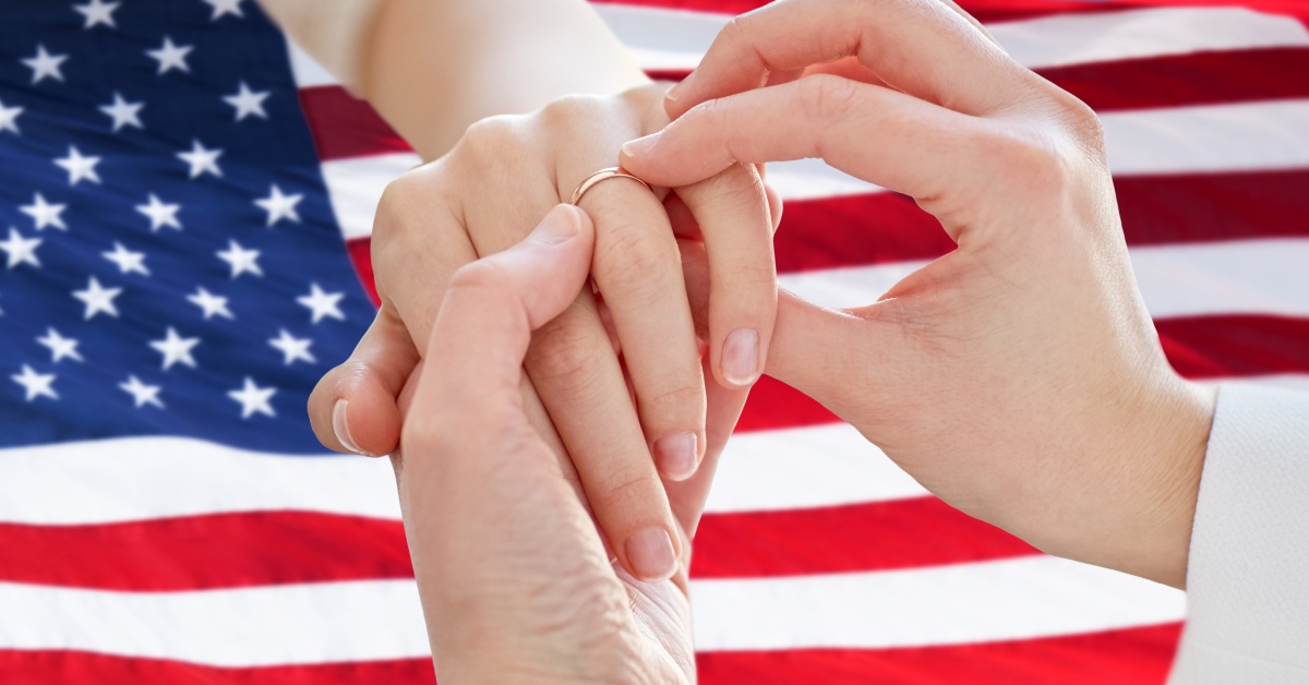 An individual placing a gold band on another person's ring finger. There is an American flag in the background.