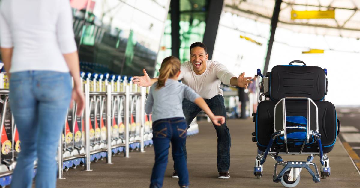 A little girl running into the open arms of an older male at an airport. The man has a luggage cart next to him.