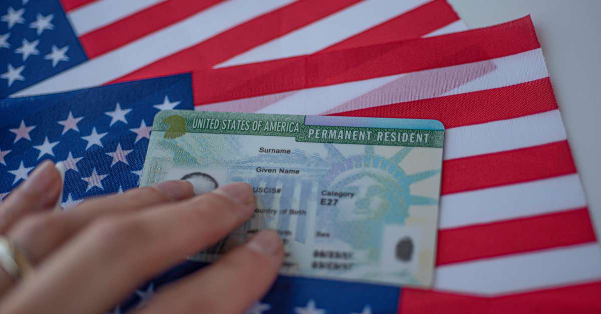 A person's hand resting on a United States of America permanent resident card. There are three US flags behind the card.