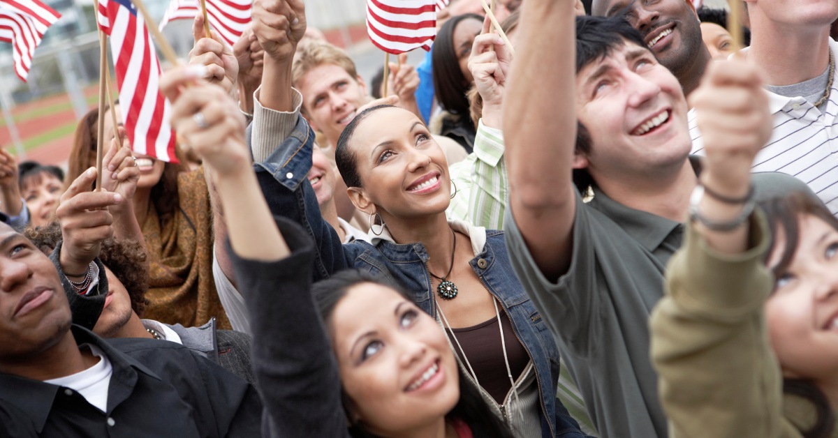 A diverse group of adults are waving small American flags. They are standing close together and looking up.