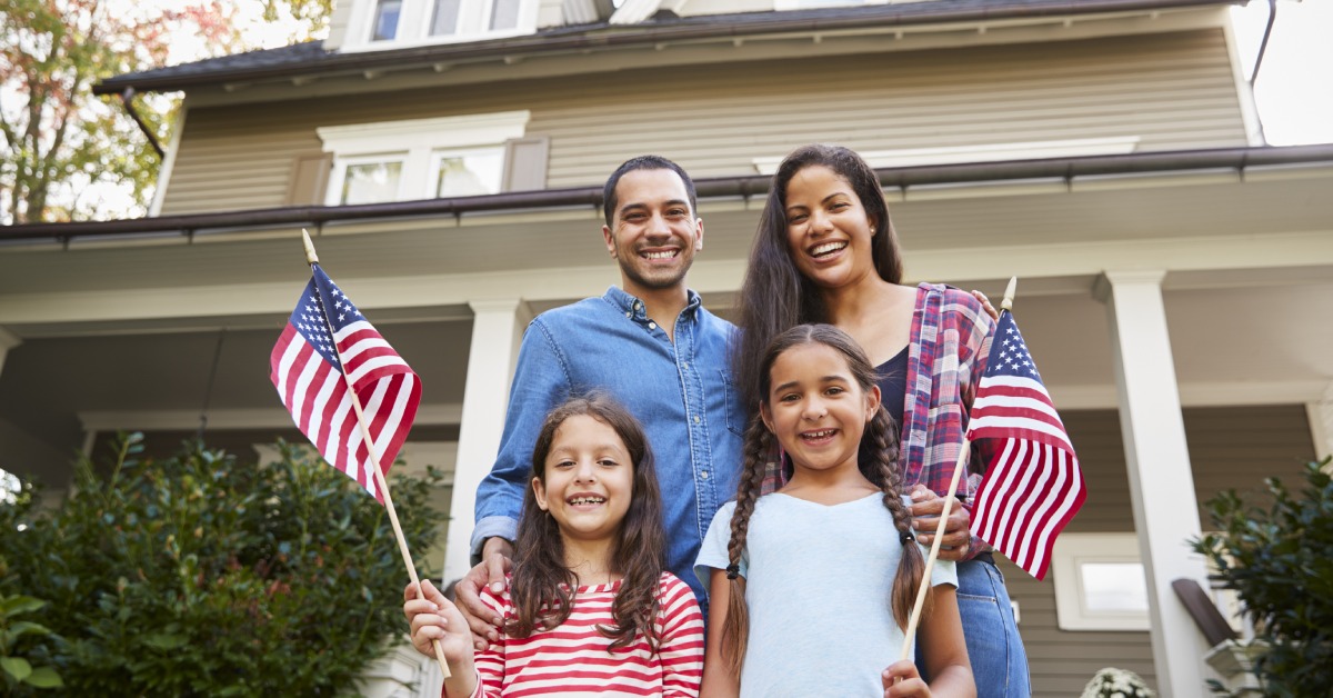 A family of four is standing in front of their house and smiling. The two children are holding small American flags.