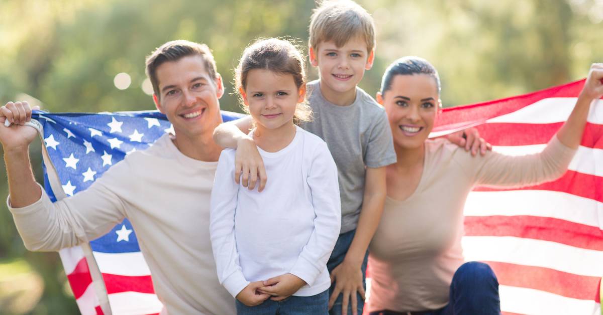 A mother and father kneeling next to their daughter and son. The adults are holding a large American flag behind them.