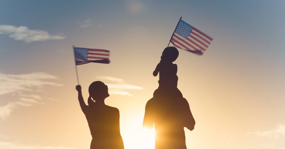 A family of three standing outside in the sunlight while waving American flags. The child is sitting on the man's shoulders.