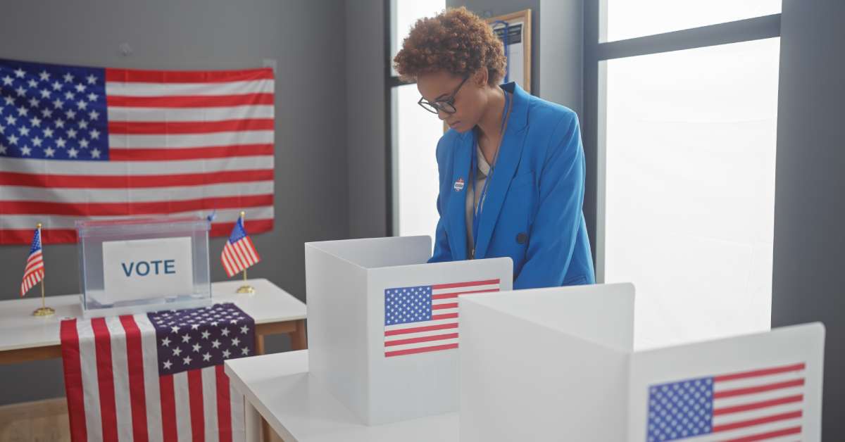 A woman in a blue blazer voting in a United States election. She is standing in a room with several American flags.