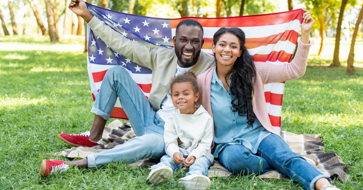 A family of three sitting on a blanket outside. They are holding a large American flag over their shoulders and smiling.