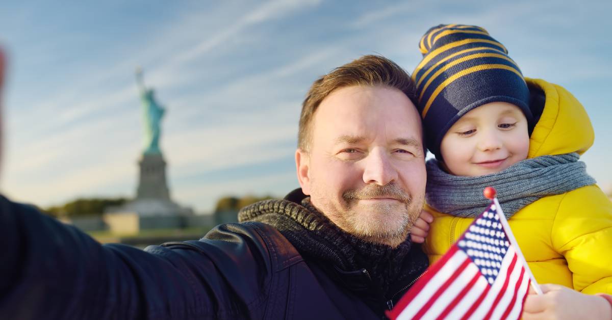 A father and son posing in front of the Statue of Liberty. The child is holding a small American flag.