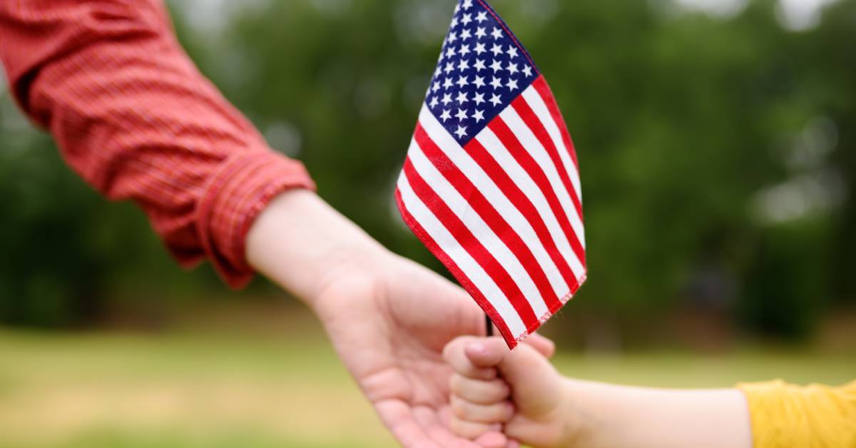 A close-up of an adult and a child holding a small American flag together. There are blurred trees in the background.