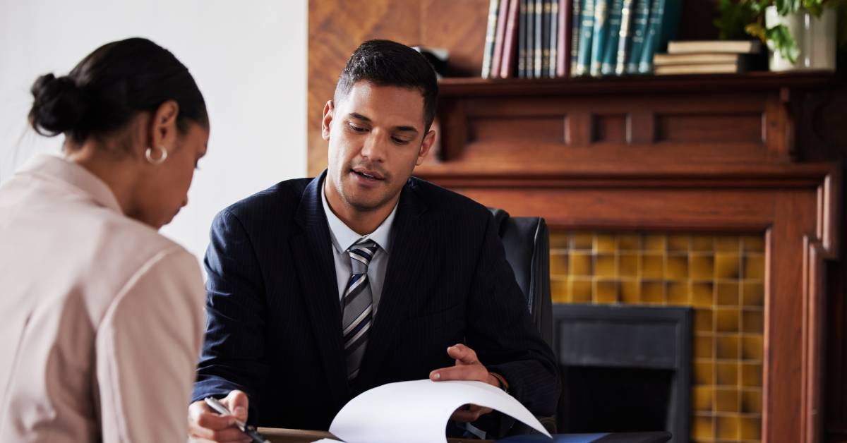 A man in a suit sitting across from a woman wearing a pink blazer. He is showing her where to sign a document.