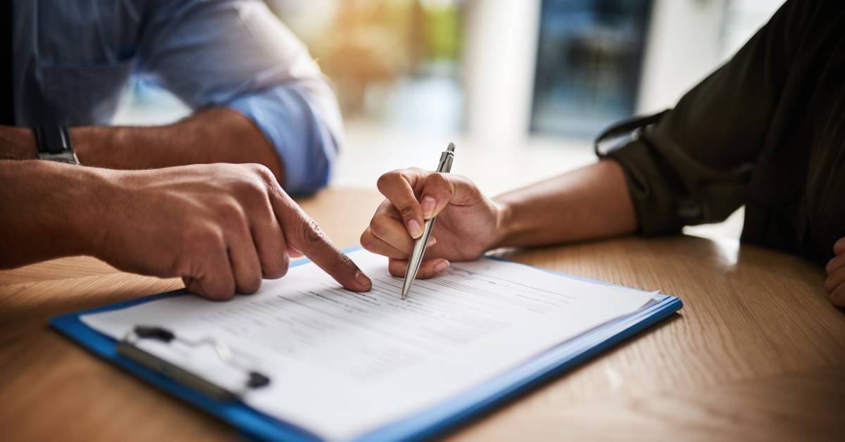 A close-up of two people sitting across from each other at a table. One person is signing paperwork on a clipboard.