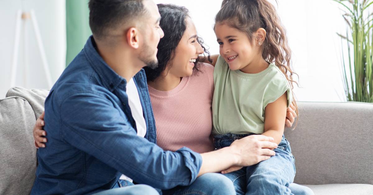 A mother, a father, and their young daughter sitting on a gray couch. They are smiling and hugging each other.