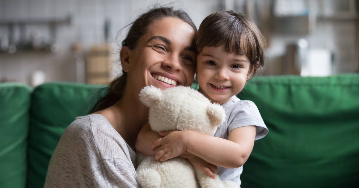 A mother and daughter smiling while hugging one another. The young girl is holding a white teddy bear.