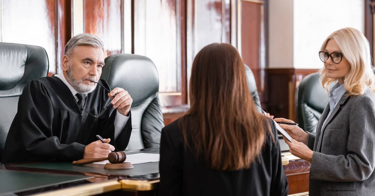 Two women dressed in professional attire standing in front of a judge in a courtroom. They are all having a discussion.