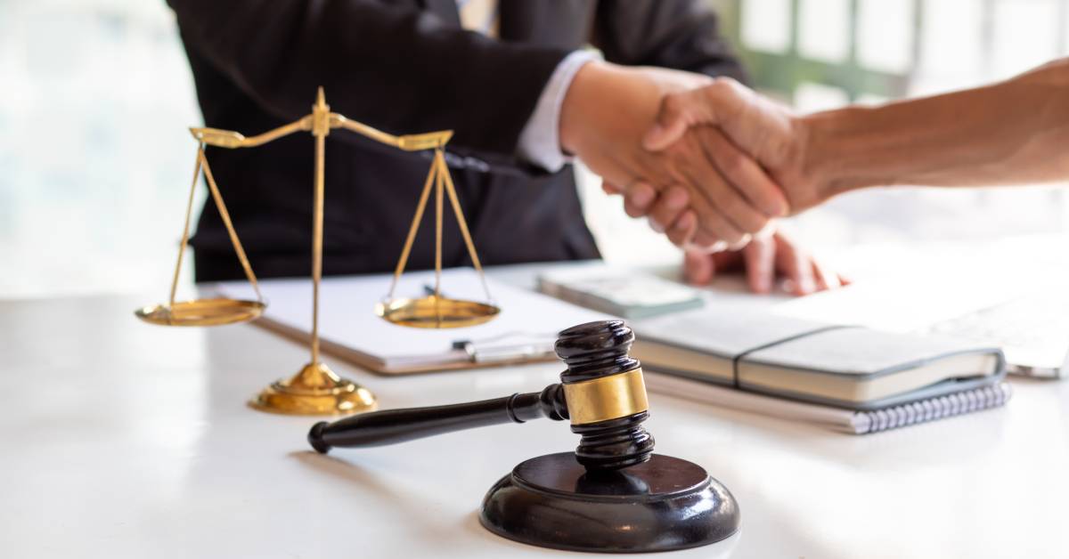 Two people sitting across from each other and shaking hands. A gavel and the scales of justice are on the table between them.