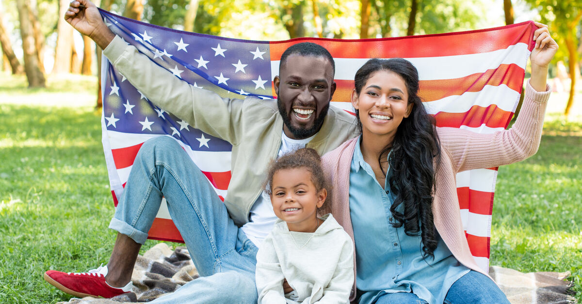 A mother, father, and daughter are sitting on the grass on a sunny day. They are holding a large American flag.