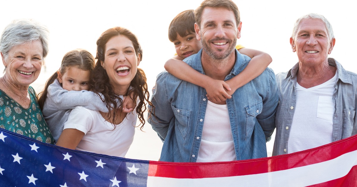 A multigenerational family is standing close to each another. They are smiling and holding a large American flag.