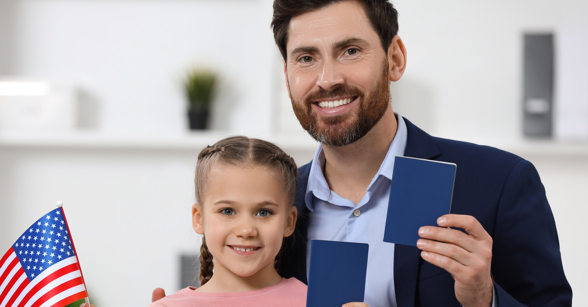 A father and daughter are smiling and holding blue passport books. The daughter is also holding a small American flag.