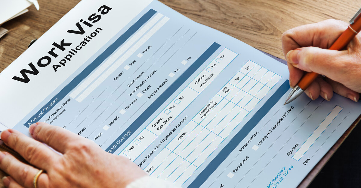 An individual wearing a sweater is holding a pen and filling out a form that says "Work Visa Application."